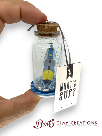 Small bottle with a sailboat inside, held by a hand, with a 'What's Sup?' tag attached, on a white background. Brand 'Bert's Clay Creations' visible.